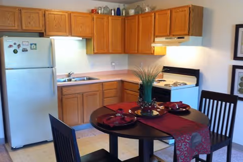 A small kitchen area with wooden cabinets, a white refrigerator, a stove with an oven, and a double sink. In front of the kitchen is a round dining table set with a red table runner, plates, and napkins, surrounded by four dark wooden chairs. There is a decorative plant centerpiece on the table.