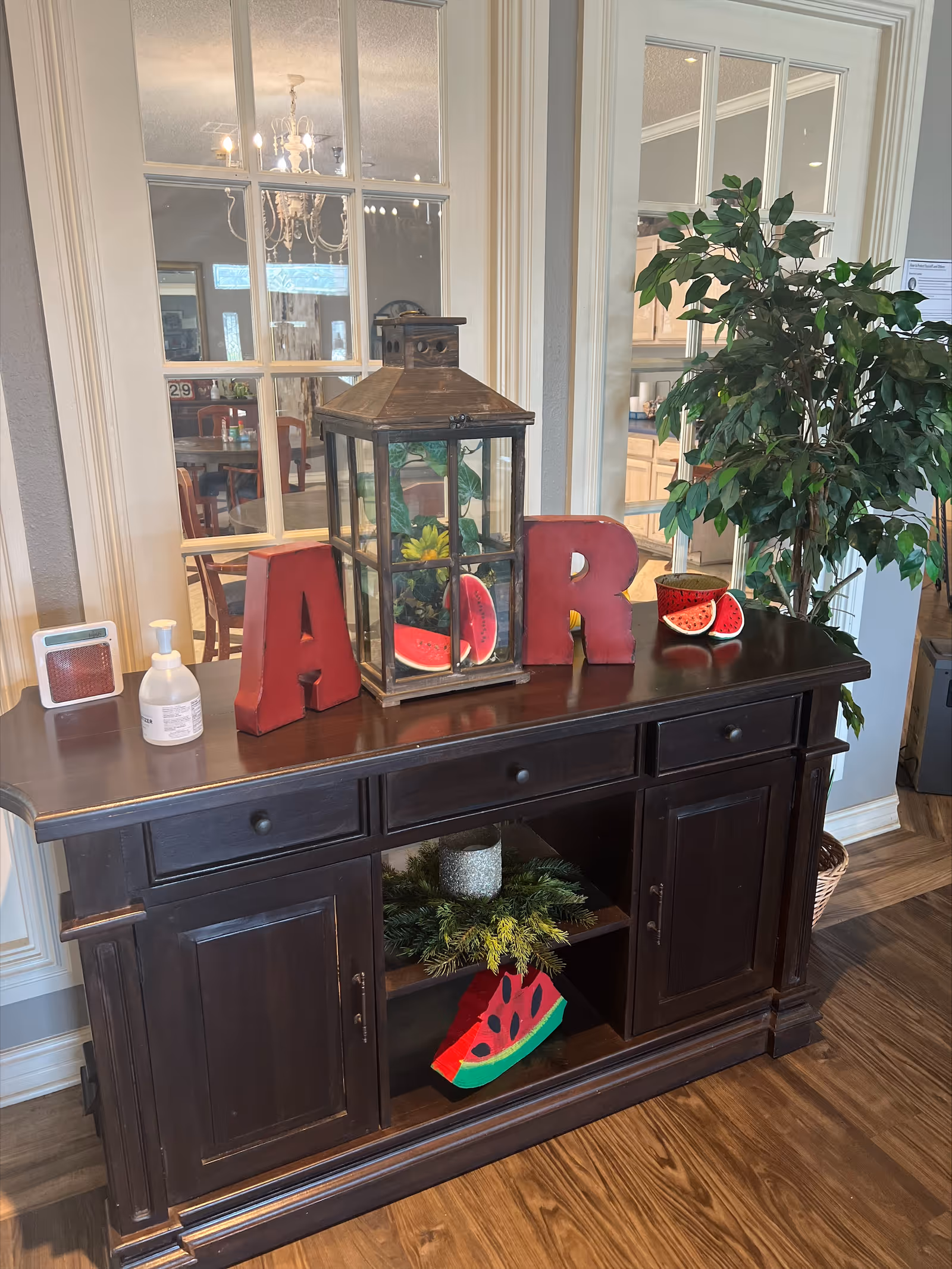 A dark wooden sideboard with decorative items on top including large red letters 'A' and 'R', a lantern with artificial sunflowers and watermelon slices inside, a small bottle of hand sanitizer, and a small speaker. There is a large green potted plant to the right of the sideboard. Behind the sideboard are glass-paneled double doors reflecting a dining area with a chandelier and wooden chairs.