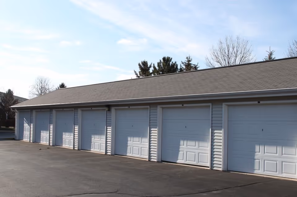 A row of seven white garage doors attached to a single-story building with a gray shingled roof, set against a partly cloudy sky with some trees in the background.
