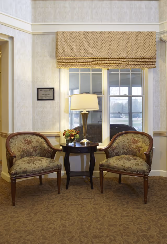 Two upholstered armchairs with floral patterns are positioned on either side of a small dark wooden table with a lamp and a small flower arrangement. Behind the table is a window with a patterned beige valance, and the walls have a light floral wallpaper with a chair rail.