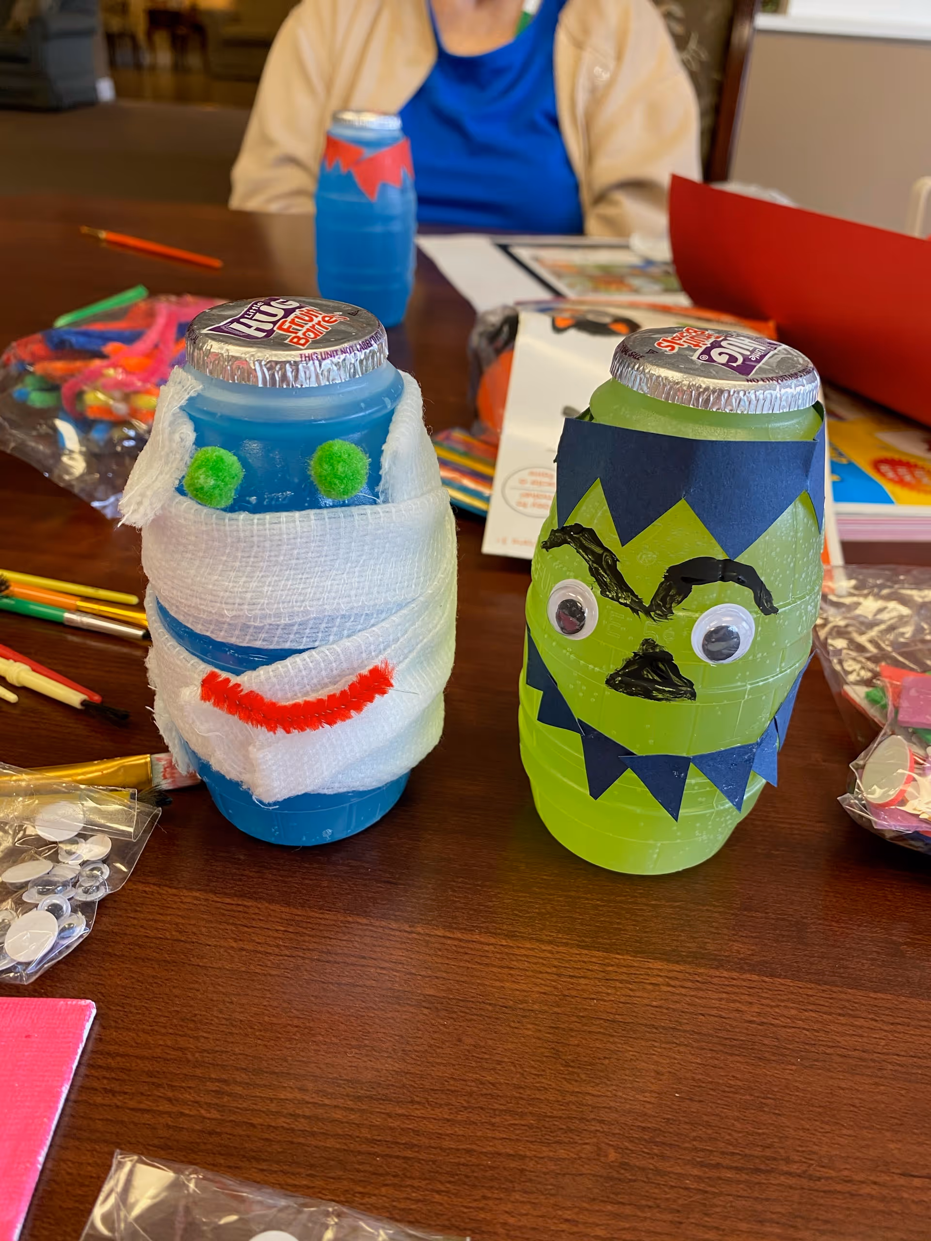 Two decorated blue and green plastic bottles on a wooden table, designed to look like Halloween characters. One bottle is wrapped in white gauze with green pom-poms for eyes and a red pipe cleaner for a mouth, resembling a mummy. The other bottle is decorated with black paper cutouts and googly eyes to look like a monster. Craft supplies and a person wearing a blue shirt and beige jacket are visible in the background.