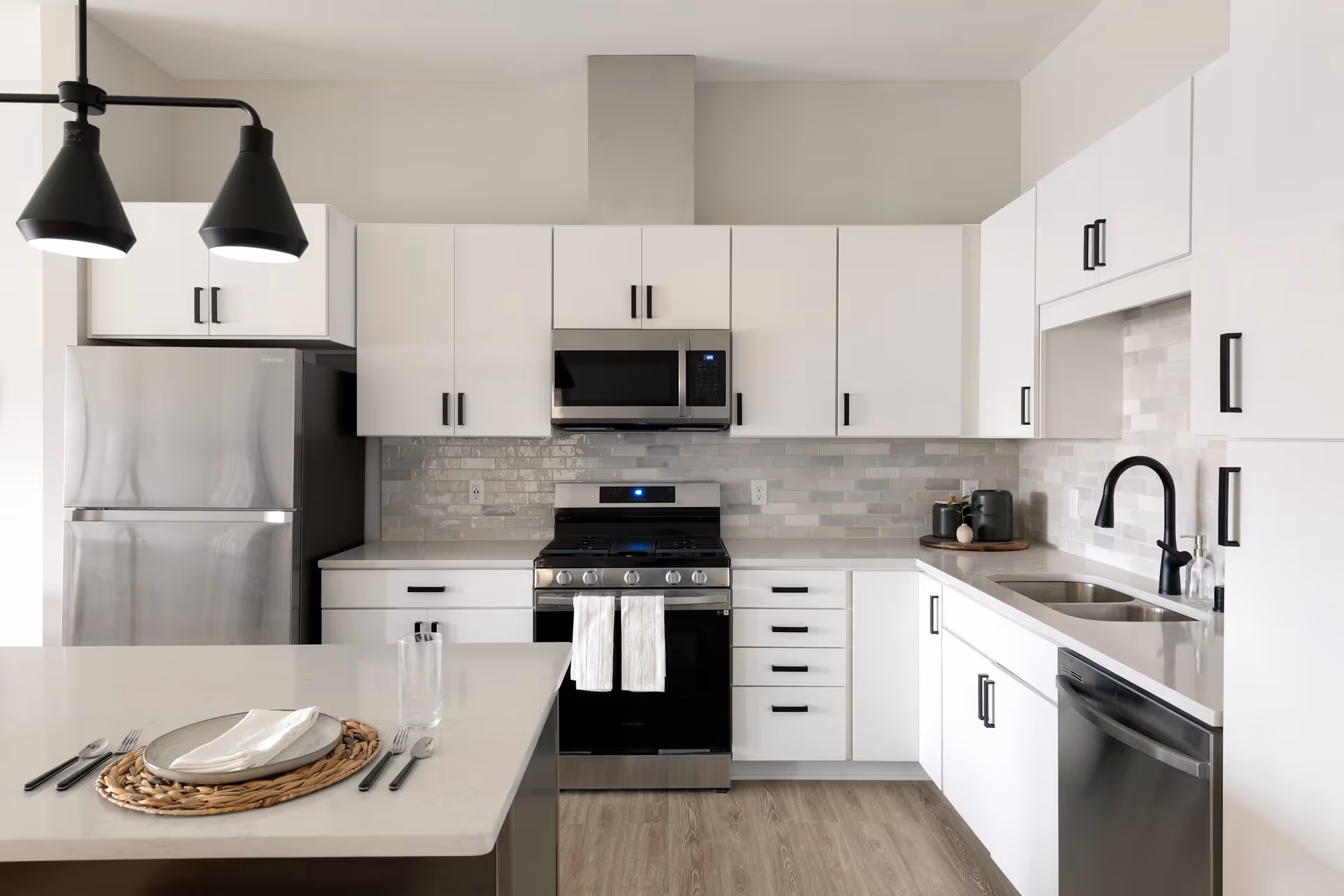 Modern kitchen with white cabinets, stainless steel refrigerator, stove, microwave, and dishwasher. The kitchen features a light gray tiled backsplash, black handles on cabinets, a black faucet, and a kitchen island set with a plate, napkin, glass, and cutlery. Two black pendant lights hang above the island.