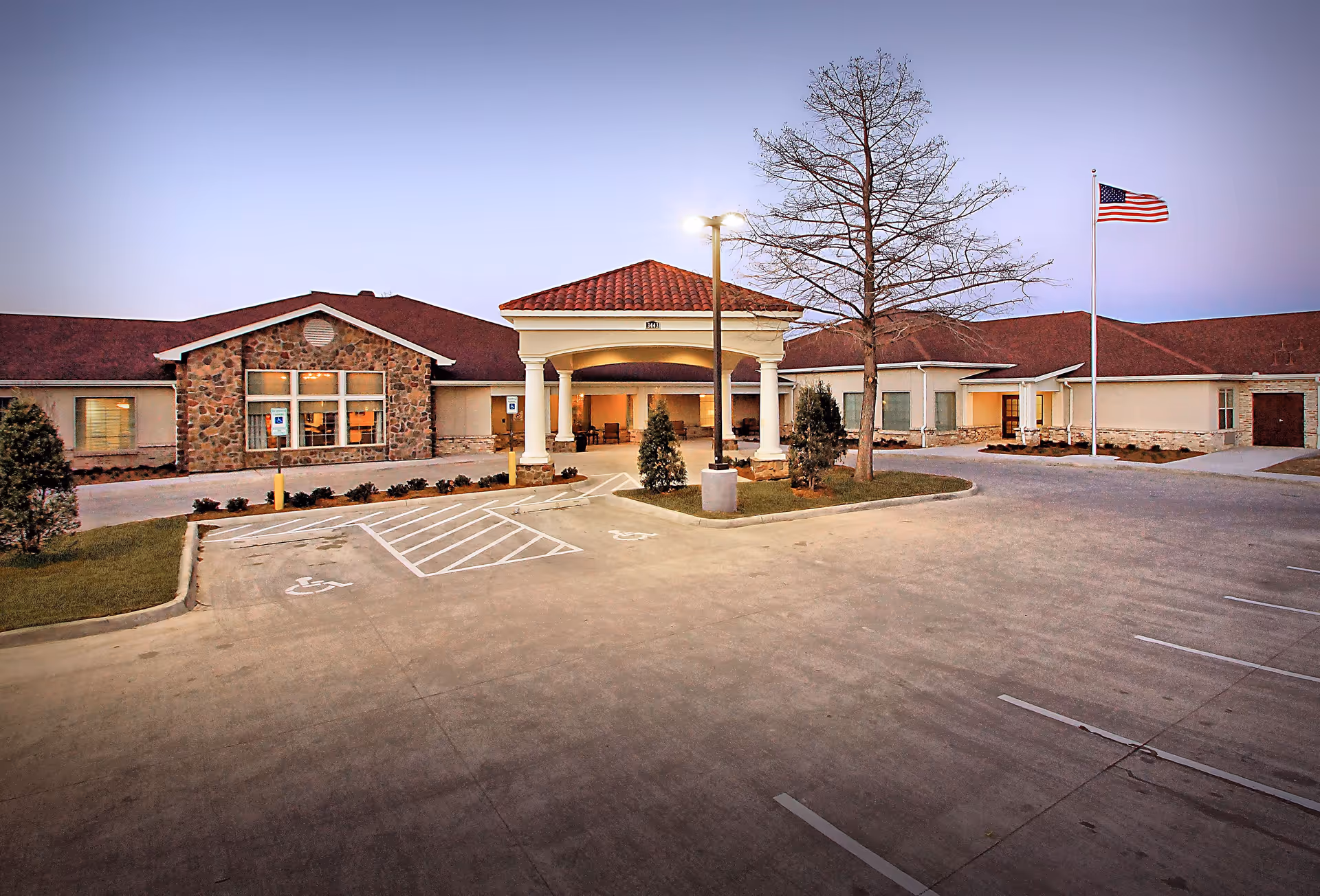 Front exterior of a single-story senior living facility with a covered porte-cochère entrance, flagpole, and empty parking lot with handicap spaces.