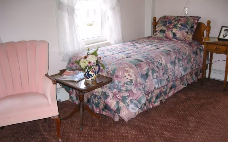 A small bedroom with a single bed covered in a floral patterned comforter and matching pillow. Next to the bed is a wooden side table with a framed photo and a small lamp. A wooden table with a vase of flowers and some magazines is placed beside a pink upholstered armchair. A window with sheer white curtains is behind the bed.