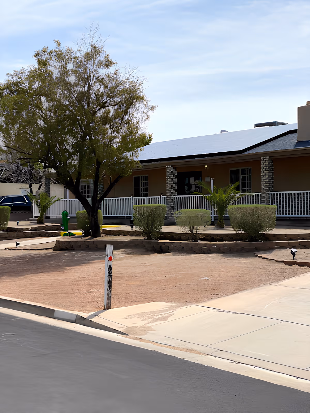 Exterior view of a single-story building with a white roof and a porch supported by brick columns. In front of the building, there is a dry, sandy yard with a few small bushes and a large tree. A paved driveway and sidewalk are visible in the foreground under a partly cloudy sky.