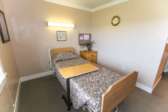 A small, simple bedroom in a senior living facility with a single bed covered in a floral patterned bedspread. There is a wooden bedside table with a plant and a bulletin board above it. A wall clock is mounted on the wall, and a rectangular light fixture is above the bed's headboard. The room has beige walls and carpeted flooring.
