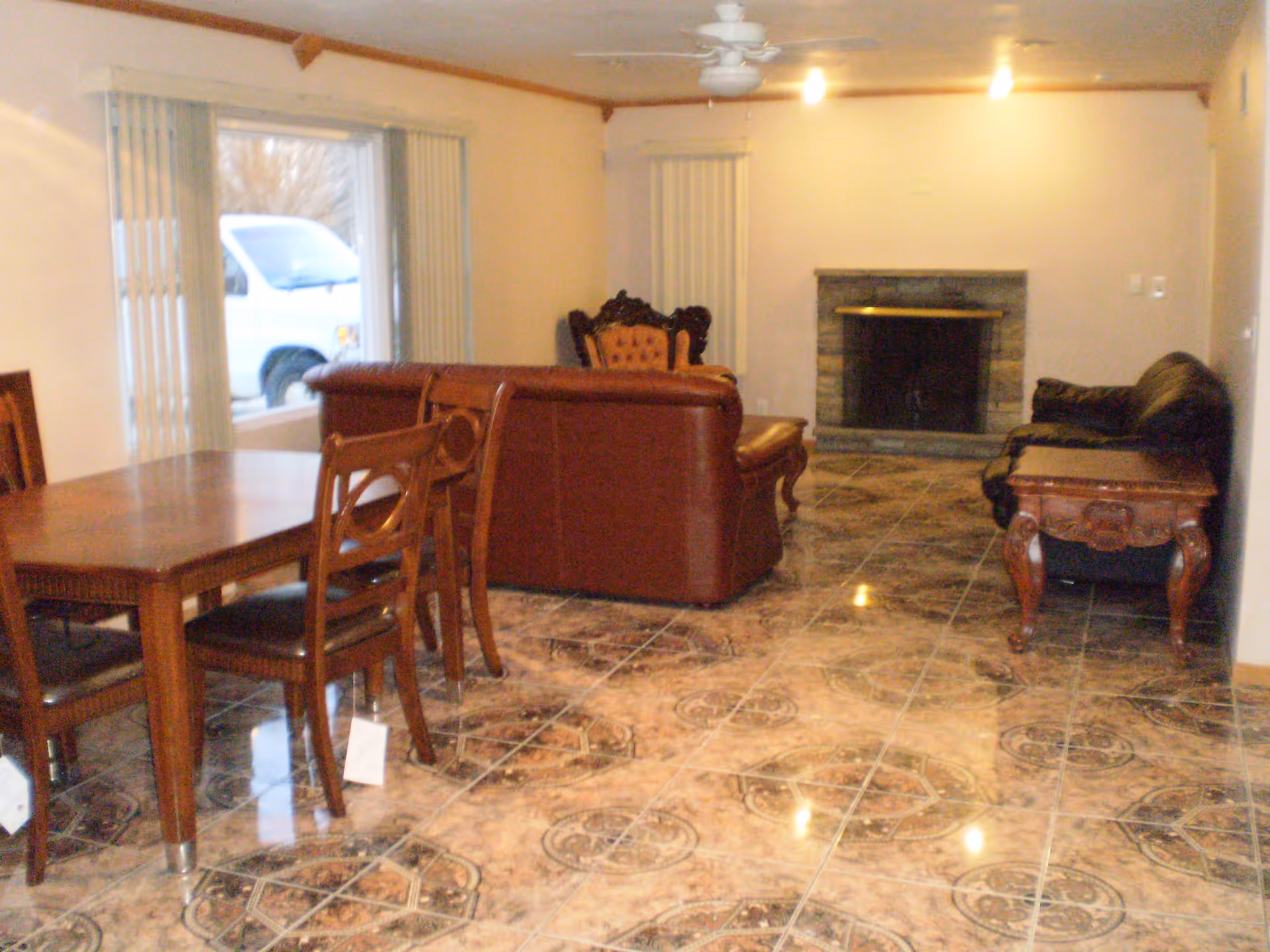 Interior view of a living room and dining area with patterned tile flooring. The room features a wooden dining table with chairs, a brown leather sofa, a black leather sofa, a wooden side table, and a stone fireplace. Vertical blinds cover the windows, and a ceiling fan with lights is mounted on the ceiling.