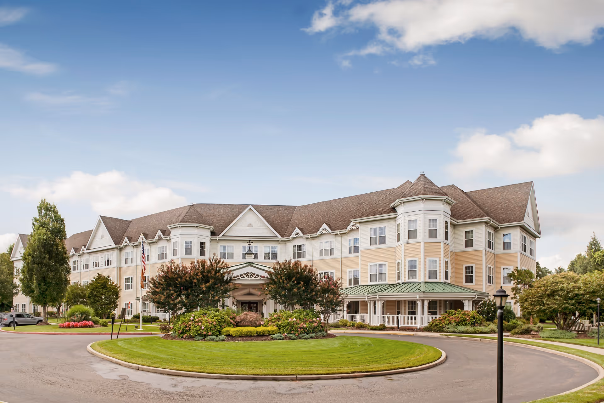 Exterior view of a large, multi-story senior living facility building with beige siding and white trim. The building has multiple windows, a peaked roof, and a covered entrance surrounded by landscaped greenery and trees. The sky is partly cloudy with blue patches.