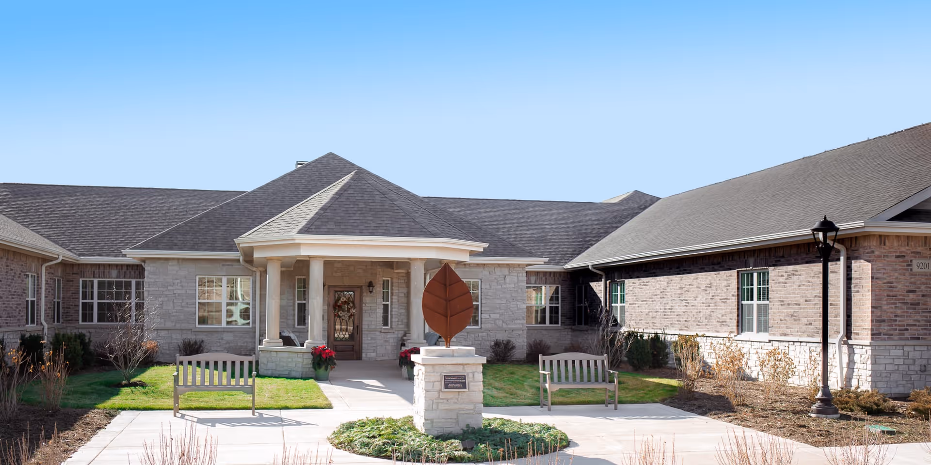 Front exterior view of Franklin Place Memory Care facility featuring a single-story building with a covered entrance supported by columns, two benches on either side of the walkway, a decorative leaf sculpture on a stone pedestal in the center, and a clear blue sky above.