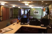 Interior view of a senior living facility reception or common area with a countertop in the foreground holding office equipment such as computers and a printer. The background shows a hallway with seating areas, green walls, and framed artwork.