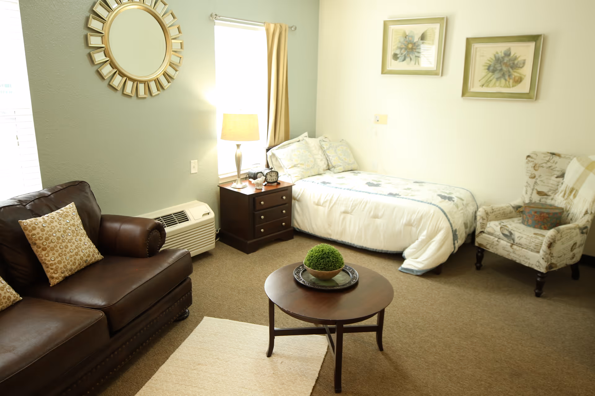 Furnished studio room with a leather sofa, coffee table, armchair, and a single bed next to a nightstand and lamp.