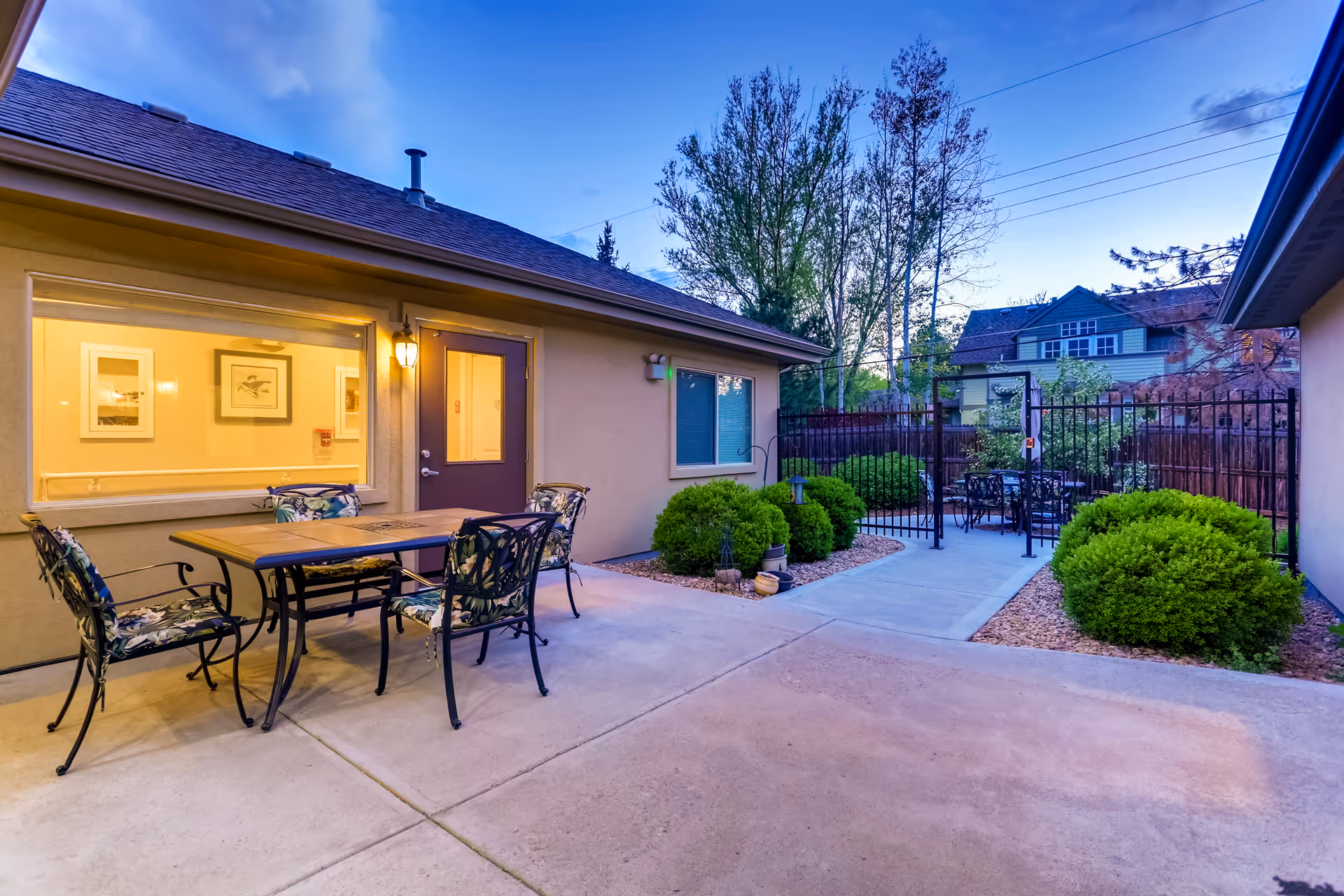 Outdoor patio area at dusk with a table and four chairs featuring floral cushions. The patio is adjacent to a building with a window and a door illuminated by a wall light. There are green bushes and trees surrounding the area, and a black metal fence with a gate enclosing another seating area with tables and chairs.