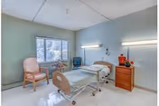 A bright care facility bedroom with a single hospital-style bed, bedside cabinet, armchair, and a window.