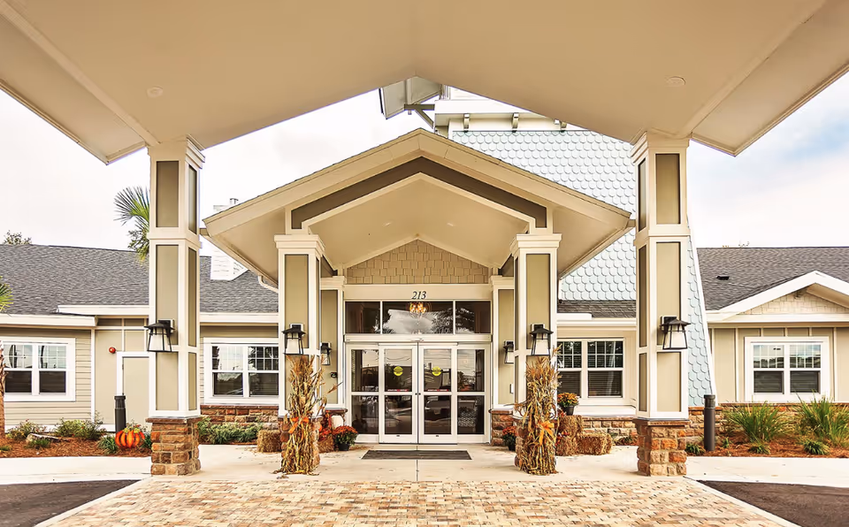 Entrance of The Canopy at Harper Lake facility with a covered driveway supported by columns decorated with autumn-themed corn stalks and pumpkins. The building has beige siding, white trim, and large glass double doors under a peaked roof.