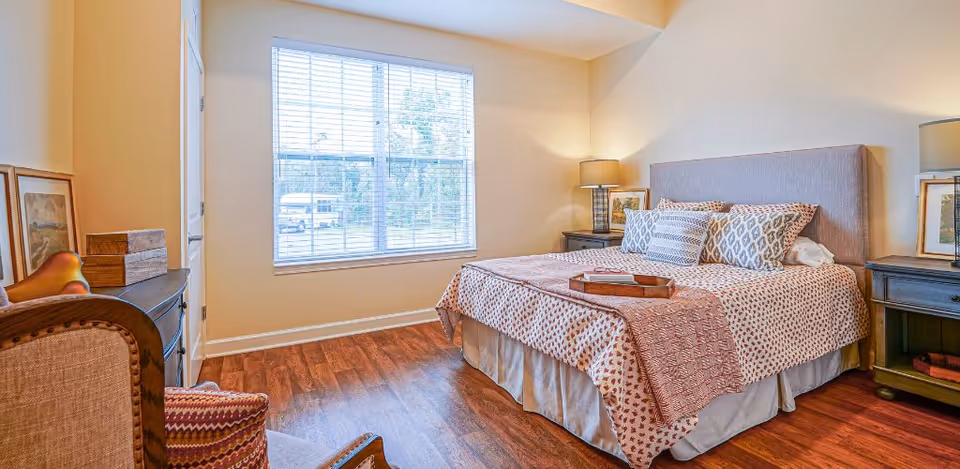 A cozy bedroom with a large bed covered in patterned bedding and multiple pillows. There are two nightstands on either side of the bed, each with a lamp and framed artwork. A window with blinds lets in natural light, and a wooden chair with a cushion is positioned near a dresser with decorative items.