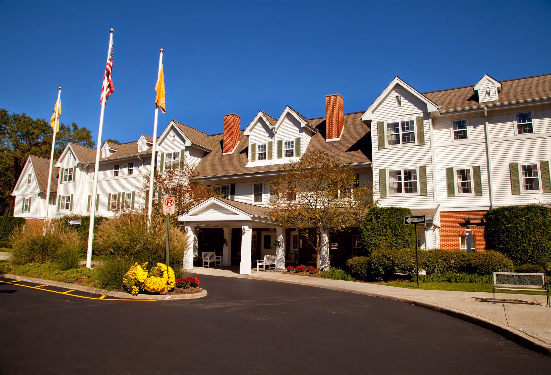 Front exterior of a multi-story white senior living facility with a covered entrance, flagpoles, and landscaped beds.