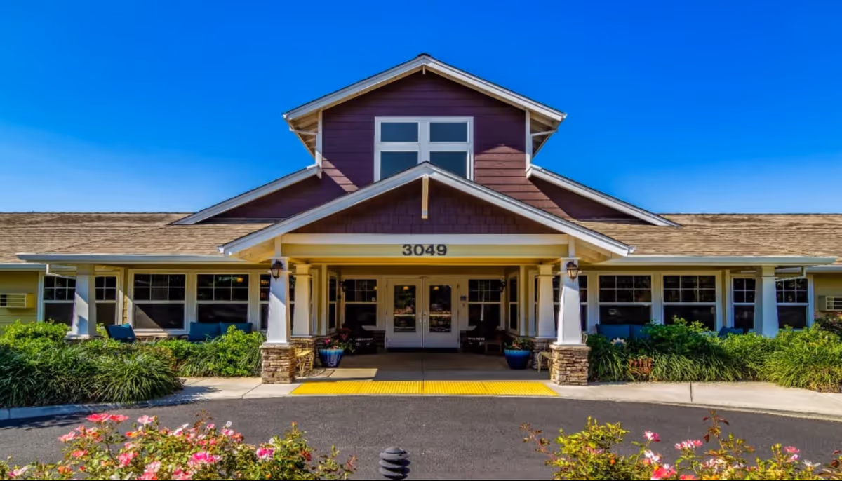 Front exterior view of a single-story building with a peaked roof and large windows. The entrance has a covered porch supported by white columns with stone bases. The building number 3049 is displayed above the entrance. There are green bushes and pink flowers in the landscaped area in front of the building under a clear blue sky.