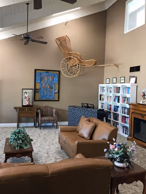 A cozy living-room style common area with leather sofas, a coffee table with plants, bookshelves, framed artwork, and a wicker rickshaw mounted high on the wall.