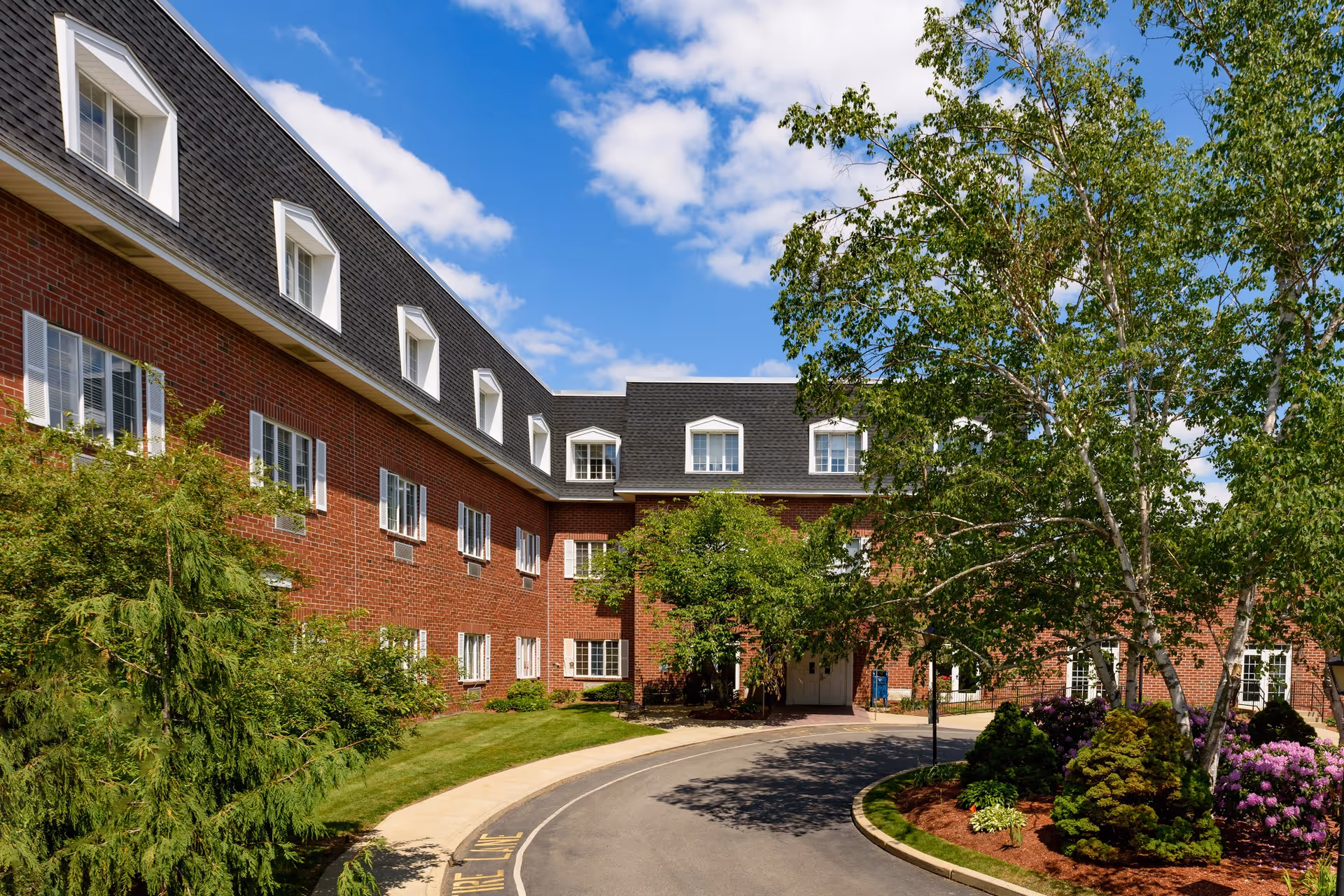 Exterior view of a brick building with multiple windows and a black shingled roof under a blue sky with some clouds. The building is surrounded by green trees, shrubs, and a curved driveway leading to an entrance.