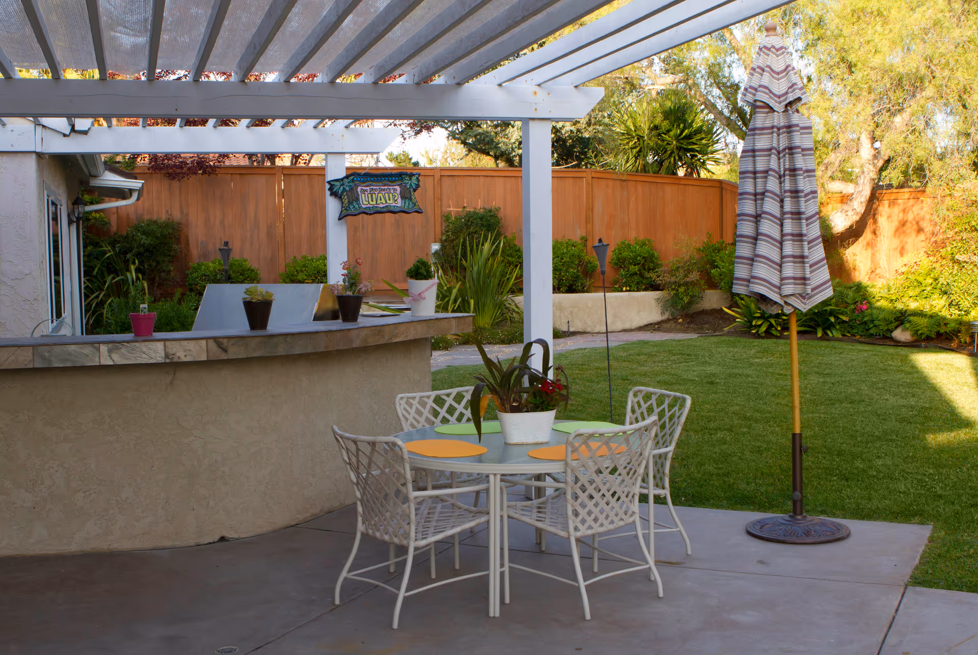 Outdoor patio area with a round glass table and four white metal chairs, each with a colorful placemat. A potted plant is on the table. There is a closed striped patio umbrella on a stand to the right. Behind the table is a curved bar counter with several potted plants on top. The area is shaded by a white pergola, and there is a wooden fence and green garden with shrubs and trees in the background. A small sign reading 'Luau' is hanging on the pergola.