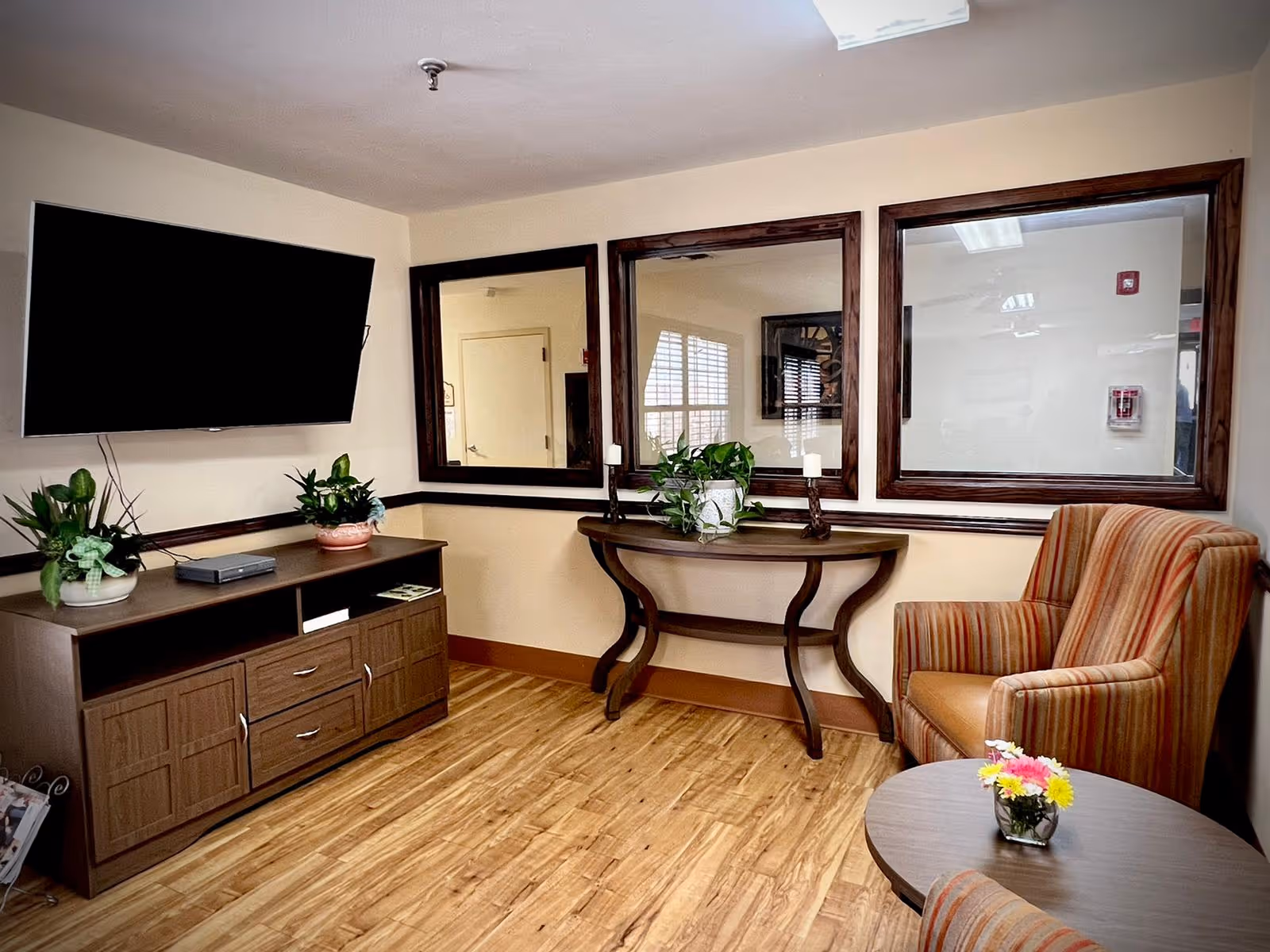 A cozy sitting area in a senior living facility with a wall-mounted flat-screen TV above a wooden cabinet holding two potted plants and a device. There is a wooden console table with a plant and two candle holders beneath three large framed windows. A striped upholstered armchair and a round table with a small flower arrangement are also visible. The room has light-colored walls and wood flooring.