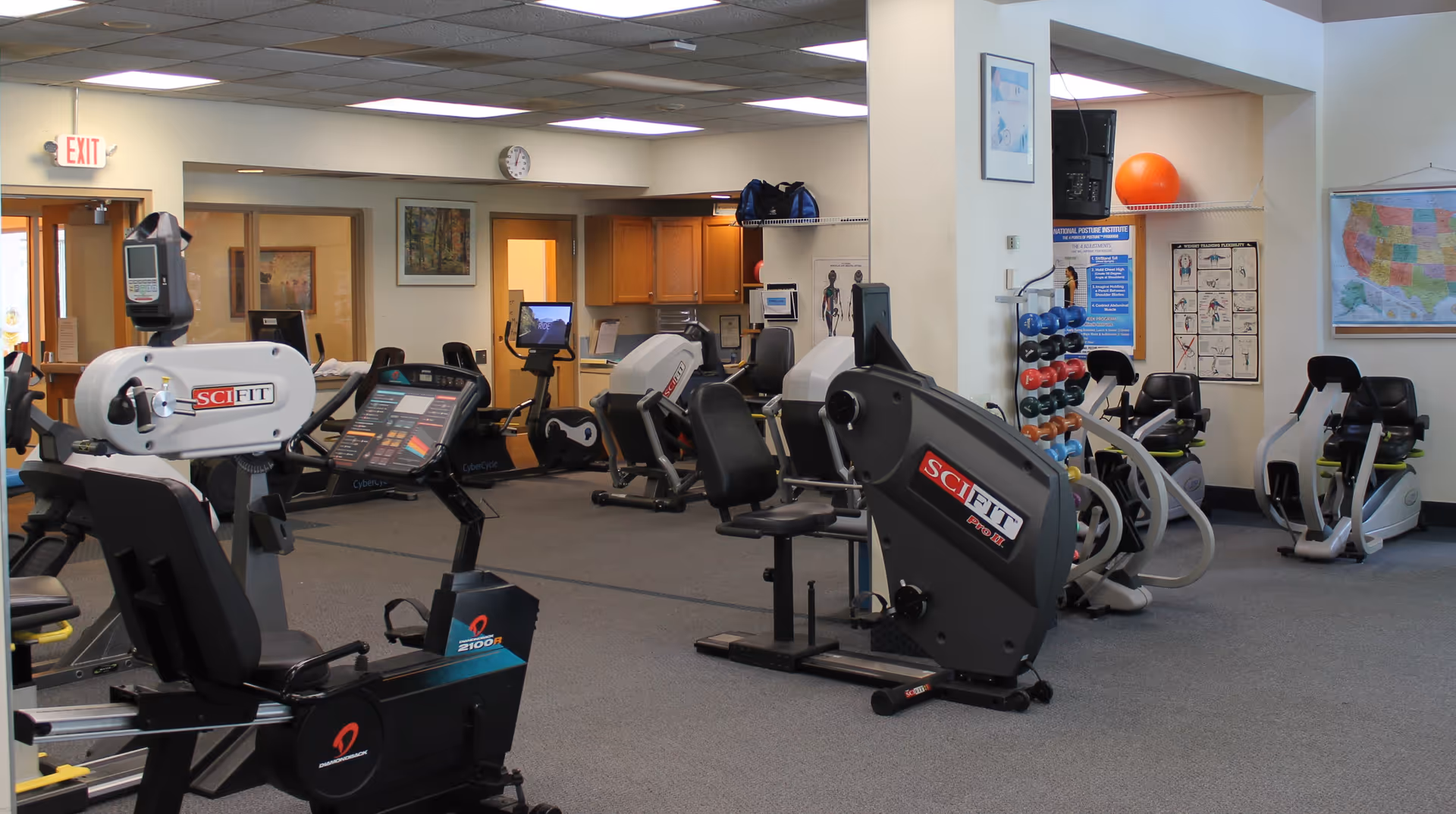 Interior view of a fitness room with various exercise equipment including recumbent bikes and weight machines. The room has carpeted flooring, white walls, and fluorescent ceiling lights. There are posters on the walls, a rack of colorful dumbbells, a wall clock, and a map of the United States. A door with an exit sign is visible in the background.
