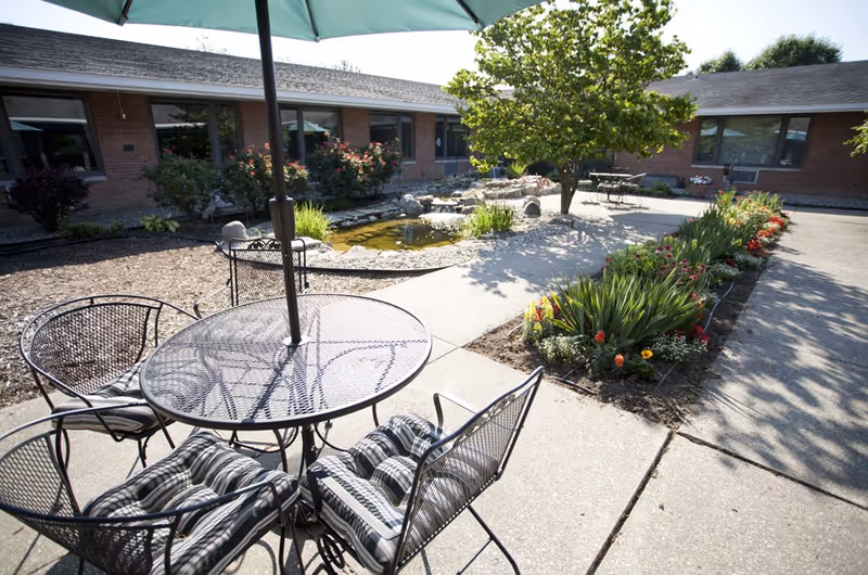 Outdoor courtyard with a metal patio table and chairs under an umbrella, flowerbeds, a small pond and a single-story brick building.