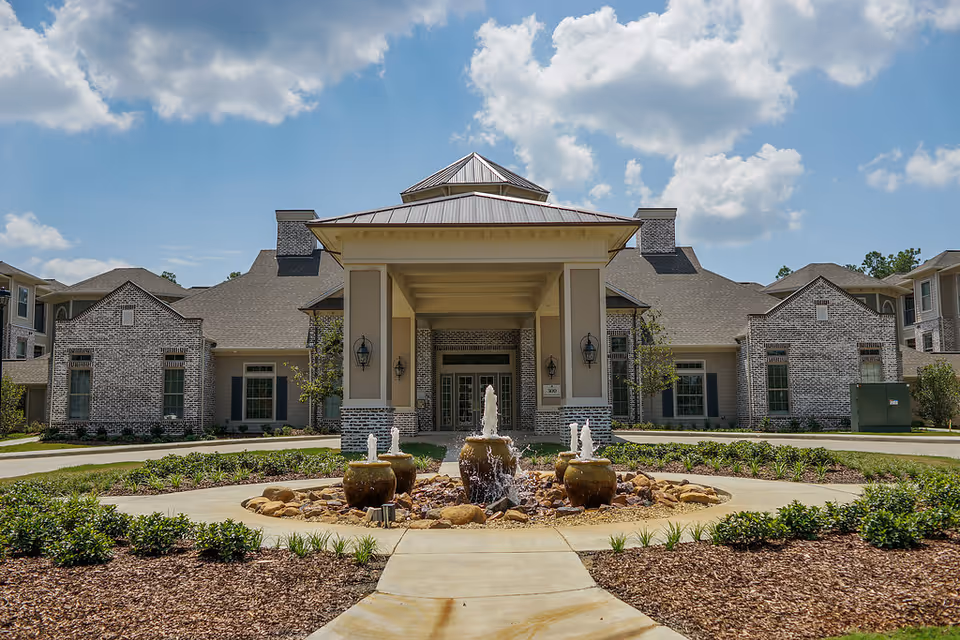 Front entrance of a senior living building with a covered porte-cochère, landscaped circular driveway and a central fountain.