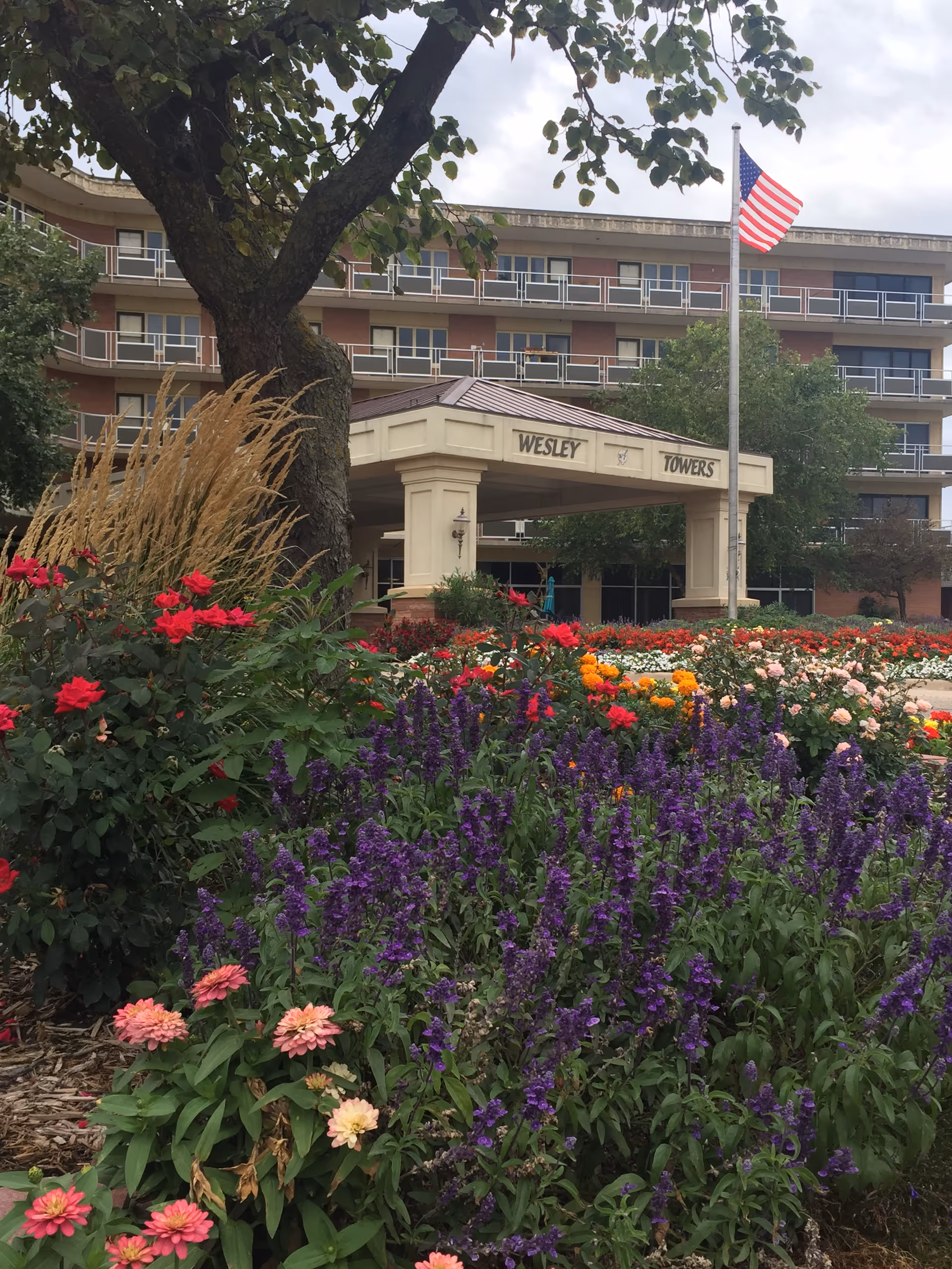 Colorful flower garden with purple, pink, red, and orange flowers in front of a multi-story building with balconies. A covered entrance with the sign 'Wesley Towers' is visible, along with an American flag on a flagpole.