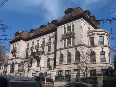 Historic multi-story stone building with arched windows and parked cars along the street.