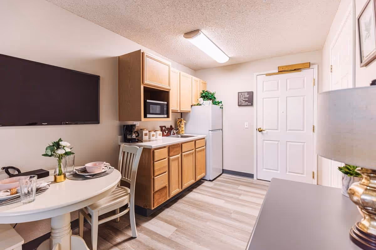 A small kitchen area in a senior living facility with light wood cabinets, a white refrigerator, a microwave, and a coffee maker. A round dining table with two chairs is set with plates, bowls, glasses, and a small vase with white flowers. A flat-screen TV is mounted on the wall above the table. The room has light-colored walls and flooring, and a white door is visible in the background.