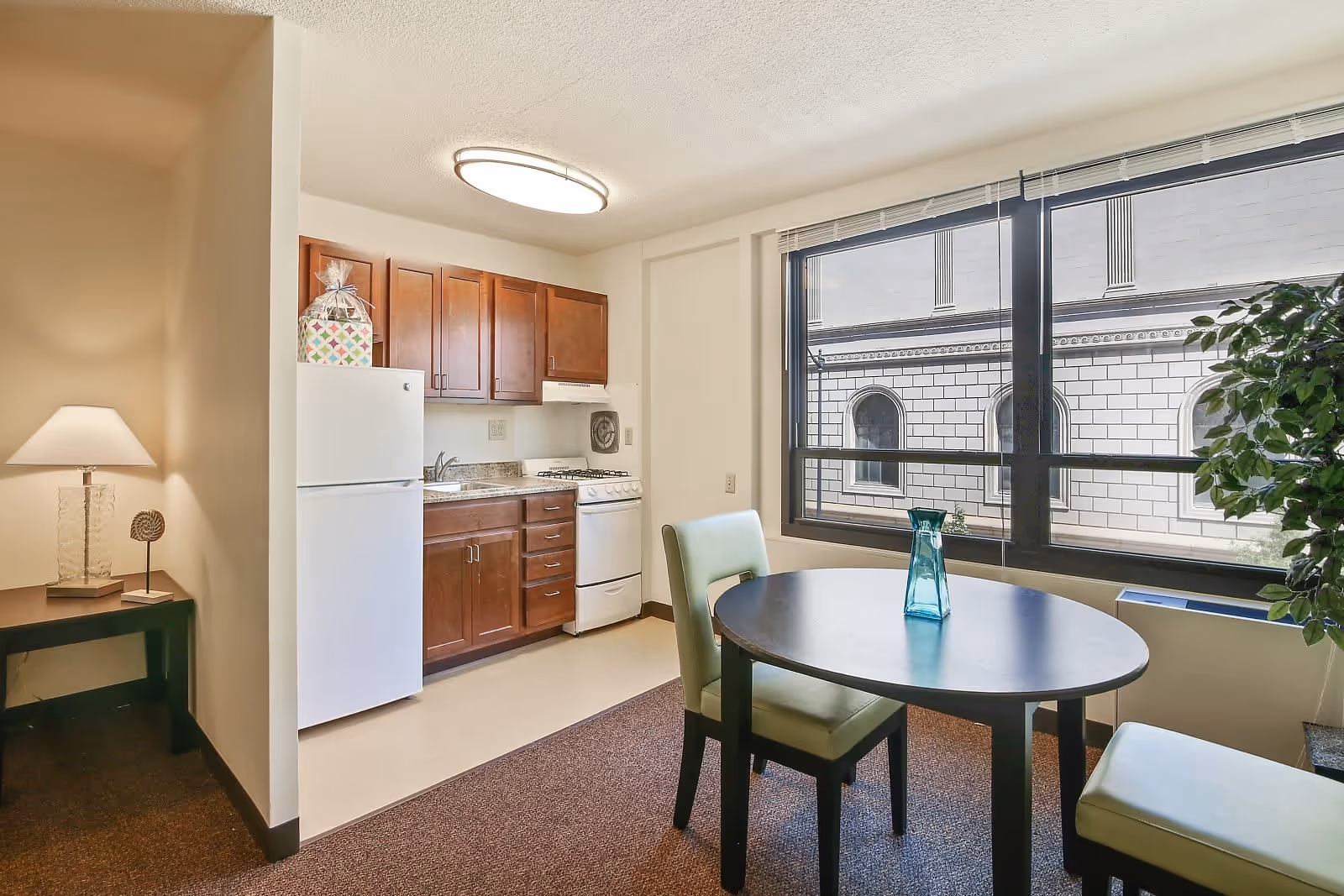 A small kitchen and dining area in a senior living apartment. The kitchen features wooden cabinets, a white refrigerator, a stove, and a sink. Adjacent to the kitchen is a round dining table with two green cushioned chairs and a blue glass vase on top. A large window shows the exterior of a building with arched windows. A small table with a lamp and decorative item is visible to the left.