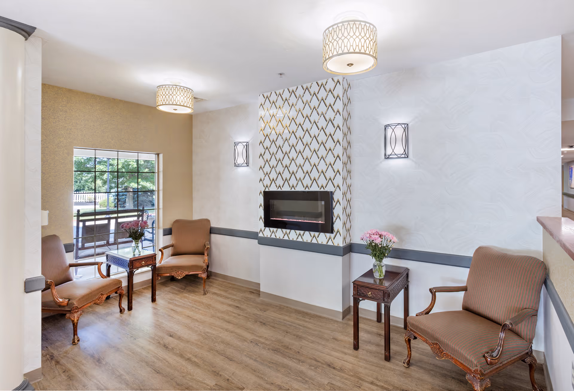 A cozy sitting area with three upholstered armchairs arranged around two small wooden side tables, each holding a vase of pink flowers. The room features a modern electric fireplace with a decorative geometric patterned wall above it. Two ceiling lights with patterned lampshades and two wall sconces provide soft lighting. A large window offers a view of greenery outside. The floor is wood, and the walls are painted in neutral tones with a chair rail molding.
