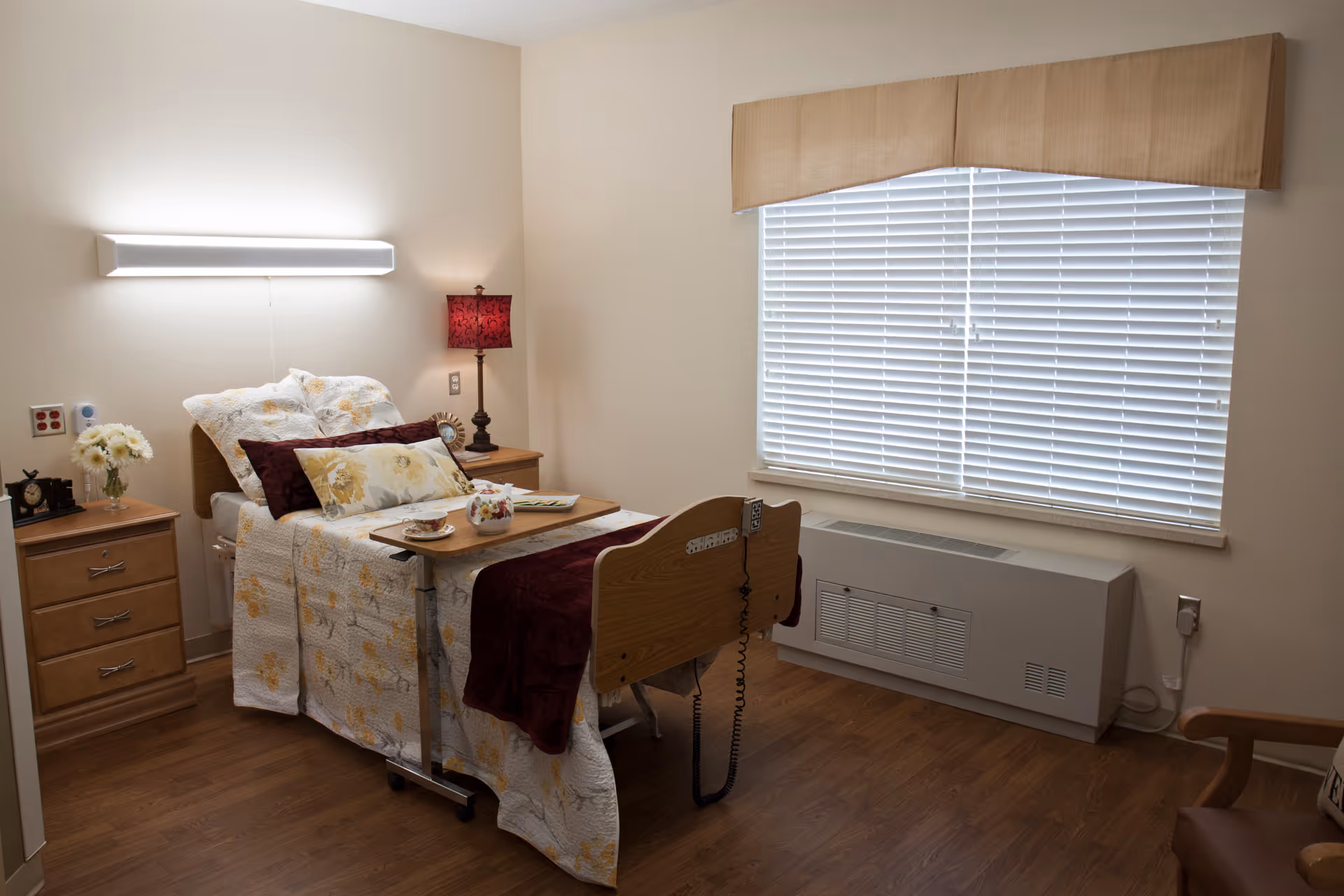 A neatly arranged bedroom in a senior living facility with a single bed covered in floral bedding and multiple pillows. There is a wooden bedside table with a lamp and a vase of flowers. A tray with a teapot and cup is placed on the bed. The room has wooden flooring, a large window with blinds, and a heating/cooling unit beneath the window.