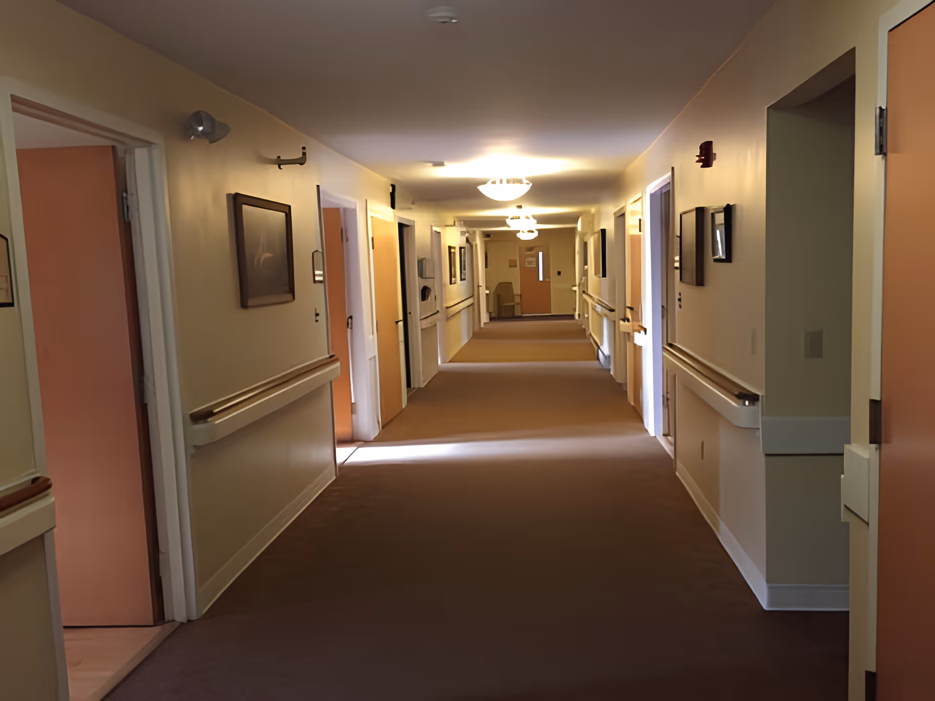 A long, well-lit hallway in a senior living facility with beige walls and carpeted floor. Several open doors lead to rooms on both sides. Handrails run along the walls, and framed pictures hang above them. Ceiling lights illuminate the corridor.