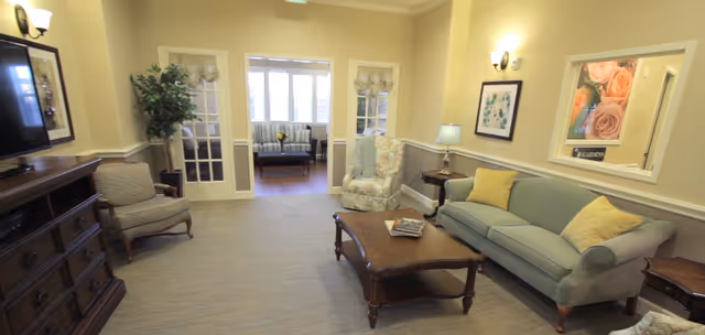 A cozy living room in a senior living facility with a green sofa, two armchairs, a wooden coffee table with magazines, a wooden dresser with a TV, framed artwork on the walls, a potted plant, and French doors leading to another room with additional seating.
