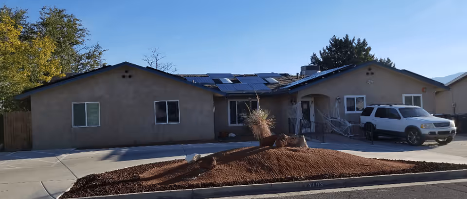 Single-story stucco building with solar panels on the roof, a white SUV parked in the driveway, and desert landscaping in front.
