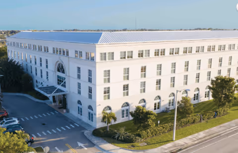 Exterior view of a large, white three-story building with many windows, surrounded by greenery and a parking area, under a clear blue sky.
