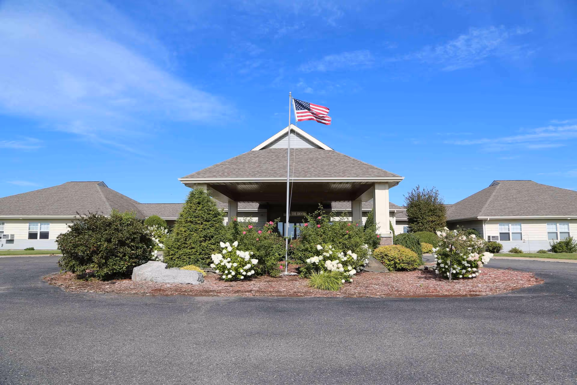 Front entrance of a single-story senior living building with a covered porte-cochere, an American flag on a pole, and landscaped shrubbery in a circular drive.
