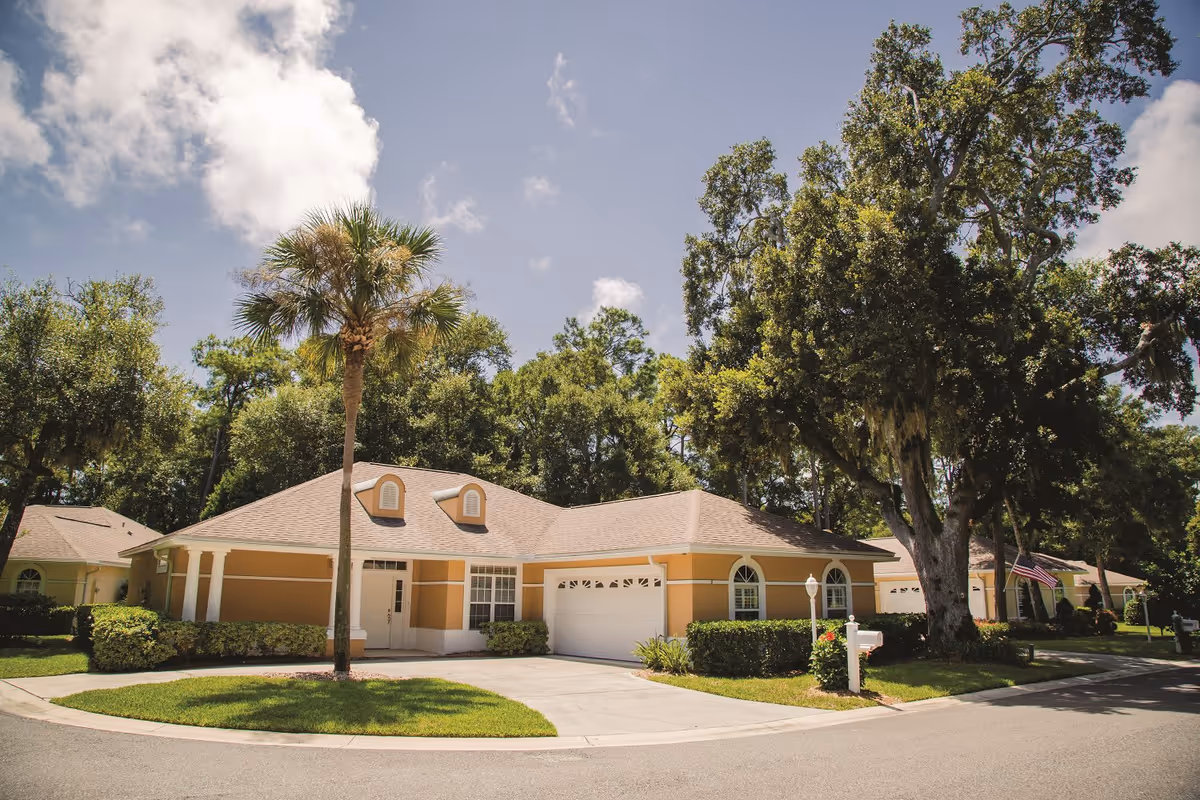 Exterior view of a single-story residential building with a beige and white facade, a garage, and a driveway. The building is surrounded by green trees, bushes, and a well-maintained lawn under a partly cloudy sky.