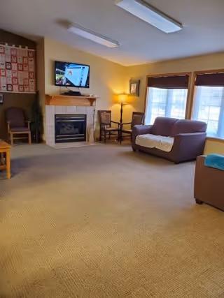 A cozy living room area with a beige carpet, a brown loveseat with a white throw blanket, a matching armchair with a blue blanket, a wooden chair, a floor lamp, and a fireplace with a mounted flat-screen TV above it. Large windows with wooden blinds allow natural light to enter the room.