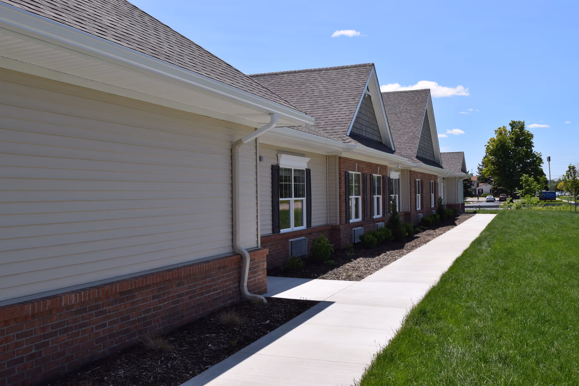Exterior side view of a single-story building with beige siding, brick accents, multiple windows with black shutters, and a gray shingled roof under a clear blue sky. A concrete walkway runs alongside the building, bordered by a mulched planting bed with small shrubs and a green lawn.