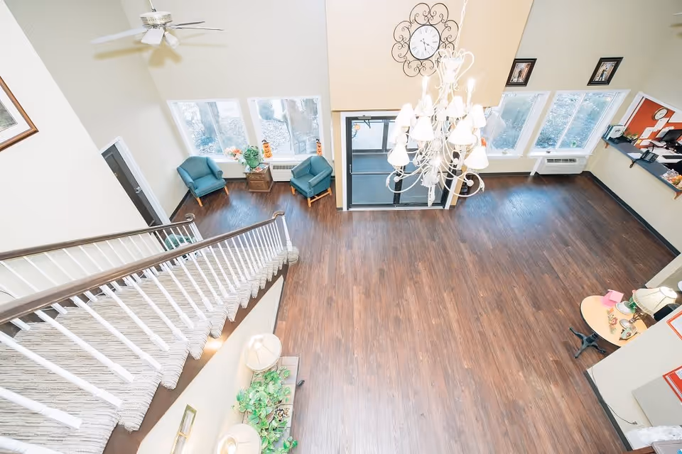 View from above of a spacious lobby area with wooden flooring, a staircase with carpeted steps and white railing, two teal armchairs near windows, a chandelier hanging from the ceiling, a clock on the wall, and a reception desk area.