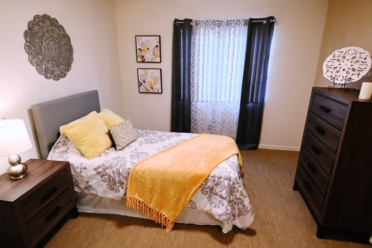 A cozy bedroom with a bed covered in a patterned gray and white bedspread, yellow and gray pillows, and a yellow throw blanket. There is a wooden nightstand with a lamp on the left side of the bed, two framed floral pictures on the wall, a window with sheer and dark curtains, and a tall wooden dresser with decorative items on top.