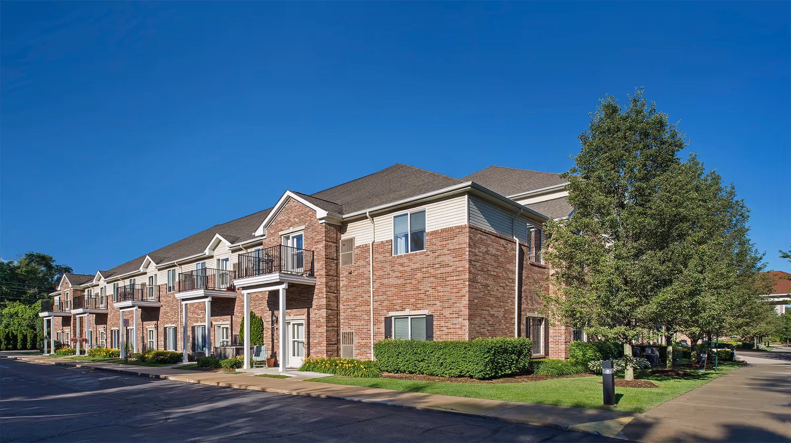 Exterior view of a two-story brick building with multiple balconies and white trim, surrounded by green bushes and trees under a clear blue sky.