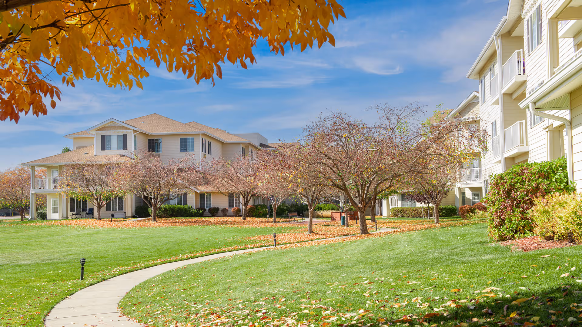A scenic outdoor view of Sugar Valley Estates | Sky Active Living featuring a curved concrete pathway through a well-maintained green lawn with trees showing autumn foliage. Beige multi-story residential buildings with balconies are visible on the right and in the background under a partly cloudy blue sky.