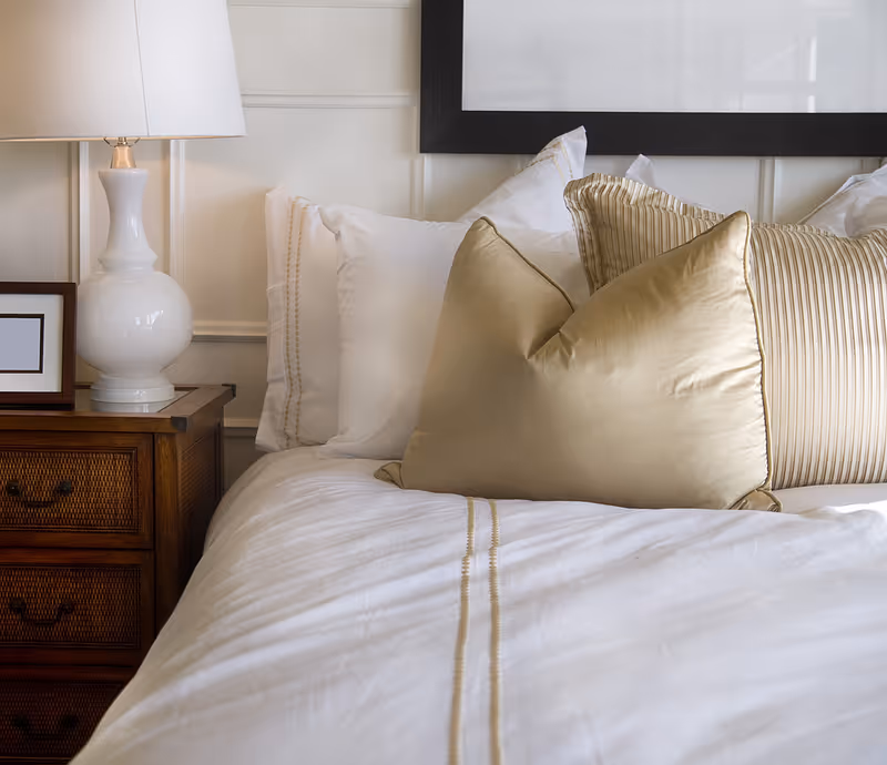 Close-up of a neatly made bed with white linens, gold decorative pillows, and a bedside table with a lamp.