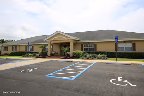Single-story building with a covered entrance, surrounded by landscaped bushes and plants. In front of the building is a parking lot with marked handicap parking spaces and signs.