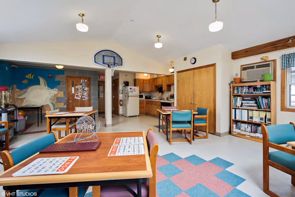 Communal activity room with tables set for bingo, chairs, a kitchenette, bookshelf, and a sea-turtle wall mural.
