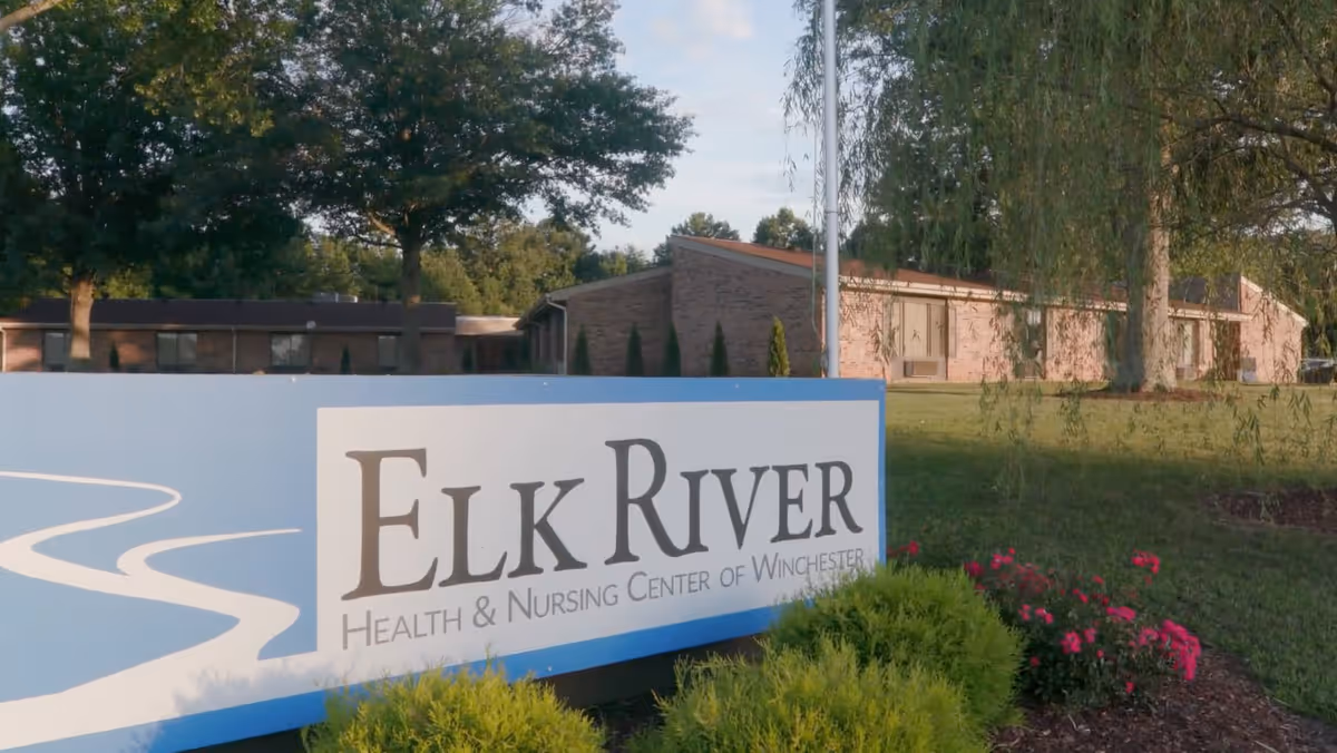 Outdoor view of Elk River Health and Nursing Center of Winchester showing a large sign with the facility name in front of a grassy area with bushes, flowers, trees, and a brick building in the background.